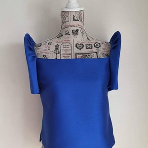May include: A vibrant blue top with unique, structured sleeves. The top is displayed on a mannequin with a vintage newspaper print design. The top has a square neckline and a tailored fit, showcasing a modern silhouette.