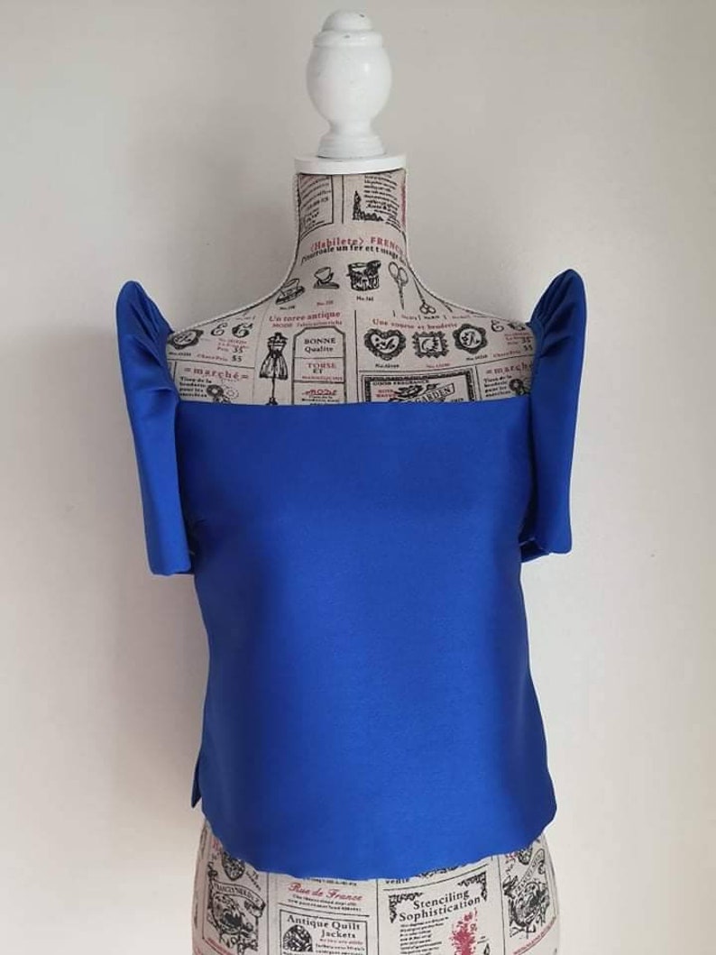 May include: A vibrant blue top with unique, structured sleeves. The top is displayed on a mannequin with a vintage newspaper print design. The top has a square neckline and a tailored fit, showcasing a modern silhouette.