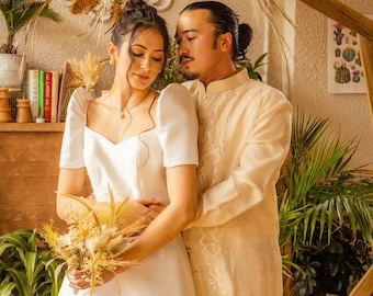 May include: A couple pose for a wedding portrait. The woman is wearing a white wedding dress with a sweetheart neckline and puff sleeves. The man is wearing a beige traditional Filipino shirt with embroidery. They are standing under a floral archway decorated with dried flowers and greenery.