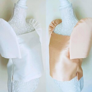 May include: Two elegant tops, one in white and the other in a soft peach color, are displayed on mannequins. Each top features a square neckline and dramatic, oversized puff sleeves. The fabric has a subtle sheen, suggesting a satin or silk material.