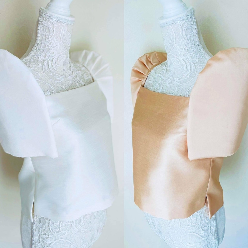 May include: Two elegant tops, one in white and the other in a soft peach color, are displayed on mannequins. Each top features a square neckline and dramatic, oversized puff sleeves. The fabric has a subtle sheen, suggesting a satin or silk material.
