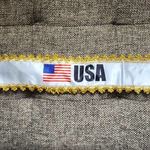 Op de afbeelding: Wit lint met gouden rand en het woord "USA" in zwarte letters. De Amerikaanse vlag is aan de linkerkant van het lint gedrukt. Het lint ligt op een getextureerd, bruin oppervlak.