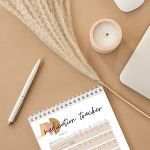 May include: A spiral-bound medication tracker notebook with the words "medication tracker" in black script. The notebook has a beige and brown abstract design. A white pen, a candle in a wooden holder, and a laptop are also visible.