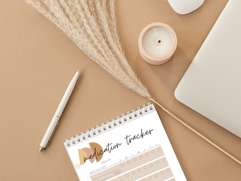 May include: A spiral-bound medication tracker notebook with the words "medication tracker" in black script. The notebook has a beige and brown abstract design. A white pen, a candle in a wooden holder, and a laptop are also visible.