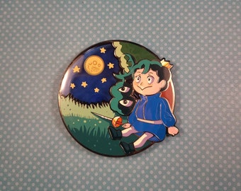 Op de afbeelding: Een ronde emaille pin met een cartoonfiguur dat een blauwe outfit en een kroon draagt, zittend op een groene heuvel met een gele maan en sterren op de achtergrond.