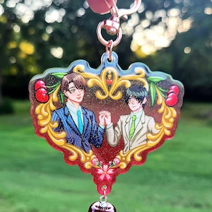 Puede incluir: Un amuleto de acrílico en forma de corazón con dos hombres con traje de chaqueta cogidos de la mano. El amuleto está decorado con filigranas doradas y cerezas rojas. Un amuleto de cereza roja cuelga de la parte inferior.