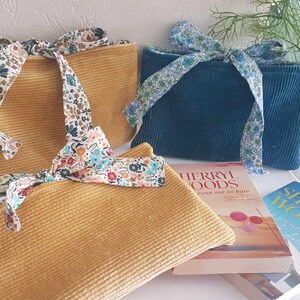Peut inclure: Trois pochettes en velours côtelé jaune moutarde et bleu sarcelle, nouées avec des rubans à motifs floraux. Les pochettes sont rectangulaires et se trouvent près de deux livres. Les livres ont le titre "Sherryl Woods" sur la couverture.