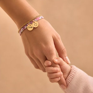 Op de afbeelding: Een armband met meerdere strengen, bestaande uit een goudkleurige metalen ketting en een roze stoffen band met bloemenprint. Twee ronde goudkleurige bedels zijn bevestigd, één gegraveerd met de naam Ambre en de andere met een zwart hartje.