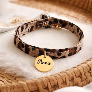 Può includere: Un collare per gatti con stampa leopardata, con un'etichetta circolare color oro. L'etichetta è incisa con il nome "Anna" in corsivo. Il collare è realizzato in tessuto fantasia e ha elementi dorati.