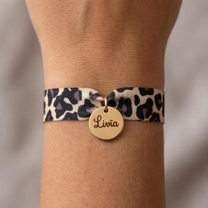 Peut inclure: Un bracelet à imprimé léopard avec un charm circulaire doré. Le charm porte le nom "Livia" gravé en écriture fine. Le bracelet est porté autour d'un poignet.
