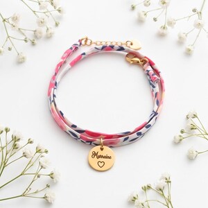 Può includere: Un braccialetto in tessuto con motivo floreale rosa, bianco e blu navy, dotato di un ciondolo circolare in metallo dorato. Il ciondolo è inciso con la parola Marraine e un piccolo simbolo a forma di cuore. Include una chiusura a moschettone dorata.