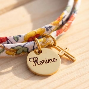 May include: A bracelet featuring a white fabric band with a colorful floral pattern in yellow, red, and green. Attached to the band are two gold-toned metal charms: a circular disc engraved with the name Florine and a small cross pendant.