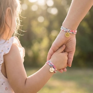 Peut inclure: Deux bracelets en tissu assortis avec des motifs floraux et des pendentifs dorés. L'un est au poignet d'un adulte, l'autre à celui d'un enfant. Les pendentifs dorés portent le nom "Zara" gravé.