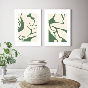 Op de afbeelding: Twee abstracte kunstprints met een groene achtergrond en witte contouren van figuren. De prints zijn wit ingelijst en hangen aan een witte muur.