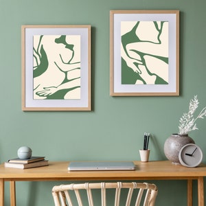 Op de afbeelding: Twee ingelijste abstracte kunstprints met groene en witte lijntekeningen van figuren, hangend aan een saliegroene muur boven een houten bureau met een laptop, een vaas met witte takken en een klok.