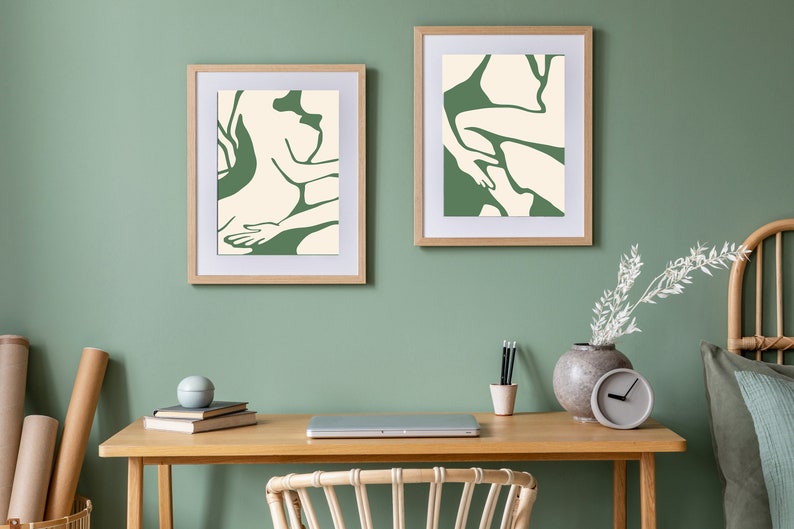 Op de afbeelding: Twee ingelijste abstracte kunstprints met groene en witte lijntekeningen van figuren, hangend aan een saliegroene muur boven een houten bureau met een laptop, een vaas met witte takken en een klok.