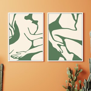 Op de afbeelding: Twee ingelijste abstracte kunstprints met een cr&egrave;mekleurige achtergrond en groene lijntekeningen van twee figuren in een liggende houding.