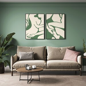 Op de afbeelding: Twee ingelijste abstracte kunstprints met groene en witte figuren op een cr&egrave;mekleurige achtergrond, hangend aan een saliegroene muur boven een beige bank met twee kussens, een salontafel en een bijzettafel met een plant.