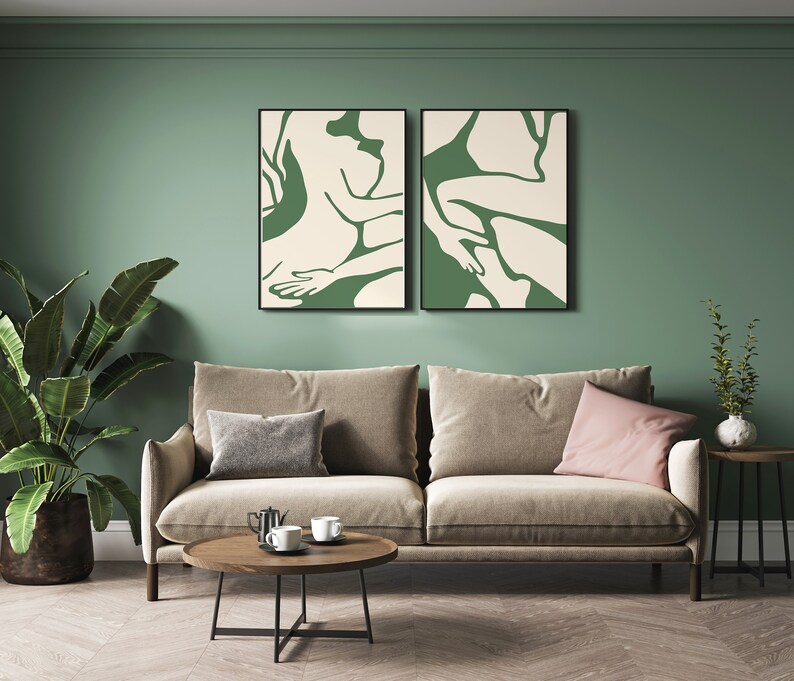 Op de afbeelding: Twee ingelijste abstracte kunstprints met groene en witte figuren op een cr&egrave;mekleurige achtergrond, hangend aan een saliegroene muur boven een beige bank met twee kussens, een salontafel en een bijzettafel met een plant.