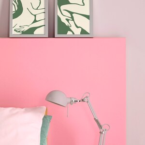 Op de afbeelding: Twee ingelijste prints van abstracte lijntekeningen in groen en wit op een roze muur boven een bed met een wit kussen en een zilveren lamp.