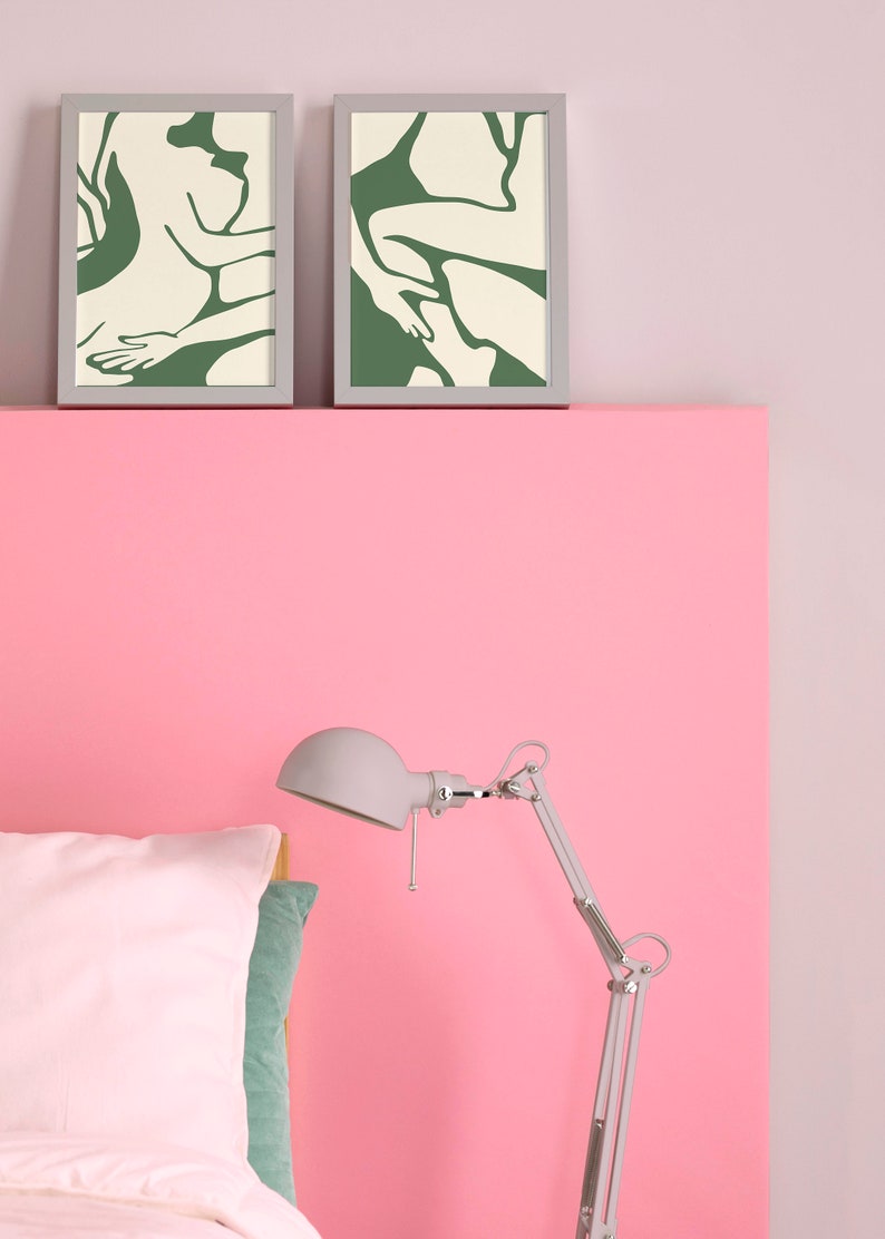 Op de afbeelding: Twee ingelijste prints van abstracte lijntekeningen in groen en wit op een roze muur boven een bed met een wit kussen en een zilveren lamp.