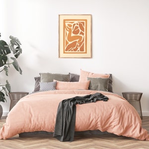 May include: A peach-colored bed with a gray throw blanket and a framed abstract art print with orange and brown shapes. The bed is in a bedroom with a large green plant and a wooden wall.