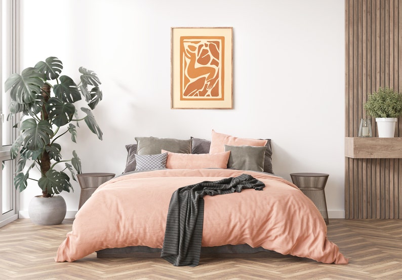 May include: A peach-colored bed with a gray throw blanket and a framed abstract art print with orange and brown shapes. The bed is in a bedroom with a large green plant and a wooden wall.