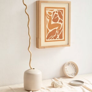 May include: A framed print of an abstract illustration of a person in shades of orange and brown on a cream background. The print is hanging on a white wall with a vase and a decorative plate on a table below.