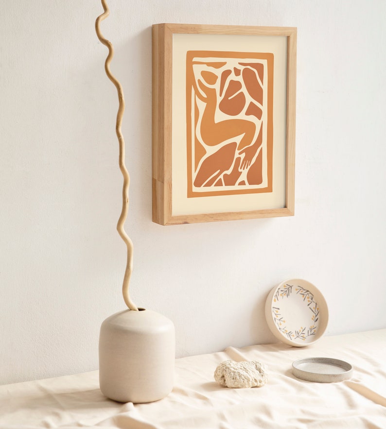 May include: A framed print of an abstract illustration of a person in shades of orange and brown on a cream background. The print is hanging on a white wall with a vase and a decorative plate on a table below.