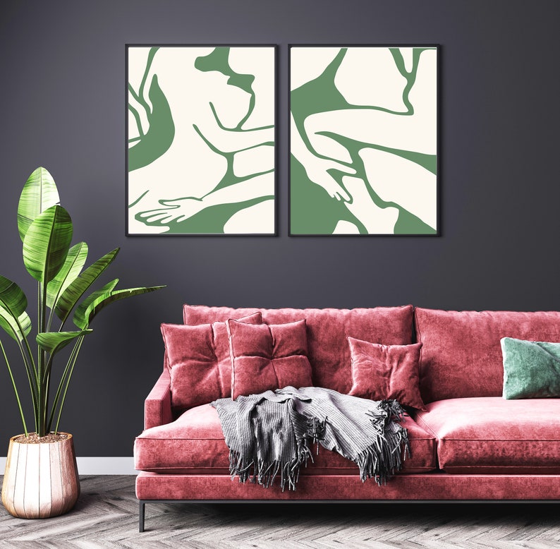 Op de afbeelding: Twee abstracte kunstprints met een groene silhouet van twee figuren op een witte achtergrond. De prints zijn zwart ingelijst en hangen aan een donkergrijze muur boven een roze fluwelen bank.