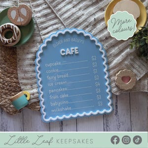 May include: A blue wooden cafe sign with a white scalloped edge, displaying a menu with items such as cupcakes and pancakes. The sign says "Oliver and Bailey's Cafe." Toy food items, including a pretzel, donut, and cookie, are placed around the sign.