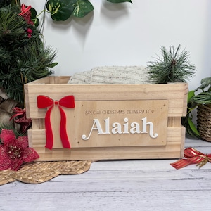 May include: Wooden crate with a red bow and the name "Alaiah" in white. The text "SPECIAL CHRISTMAS DELIVERY FOR" is above the name. The crate is filled with a light-coloured blanket and decorated with greenery.