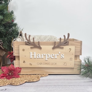 May include: A wooden Christmas box, personalised with "Harper's" in white lettering, and decorated with brown antlers. The box contains a cream blanket. The words "CHRISTMAS BOX" are below the name, with festive icons.