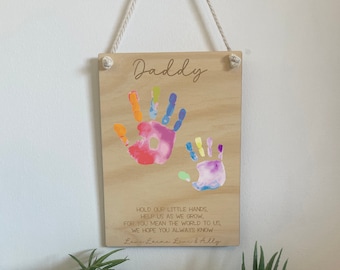 Puede incluir: Placa de madera con la palabra "Daddy" y huellas de manos coloridas. La placa incluye el texto "Hold our little hands, help us as we grow, for you mean the world to us, we hope you always know." Un artículo decorativo.