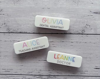 May include: Three white rectangular name badges with rounded corners. Each badge features a name in rainbow colours and a job title. The names are Olivia, Alice, and Leanne. The job titles are Dental Assistant, Teaching Assistant, and Doctor.