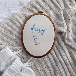 May include: An oval wooden embroidery hoop containing off-white fabric with the name daisy embroidered in blue script. Below the name is a small white goose with an orange beak and feet, wearing a blue bow tie, centred on the fabric surface.