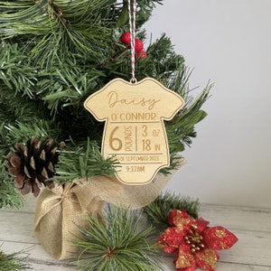 May include: A wooden baby onesie-shaped Christmas ornament. The ornament is engraved with the name "Daisy O'Connor", birth weight of 2.72 kg 85 g, and length of 45.72 cm, along with the birth date and time. The ornament hangs from a red and white string.