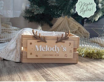 May include: A rectangular wooden crate featuring a light-colored front panel with the text "Melody's CHRISTMAS BOX" in white and brown lettering. The panel is decorated with small icons and topped with two brown reindeer antlers.