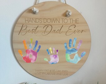 May include: Round wooden sign with the text "HANDS DOWN TO THE Best Dad Ever". The sign features colorful handprints and the text "HAPPY FATHER'S DAY 2023 LOVE RYLAN IVIE AND LEXI". It has a rope for hanging.