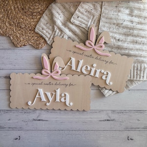 May include: Two wooden Easter signs with scalloped edges. Each sign features a bunny ear design with a pink bow. The signs read "a special easter delivery for" followed by a name in white letters. One sign says "Aleira" and the other says "Ayla".