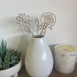 May include: A white ceramic vase displays wooden flower cut-outs, including a daisy and a rose. A small succulent plant in a white bowl and a marble-effect candle are also present. The arrangement is on a wooden surface against a white wall.