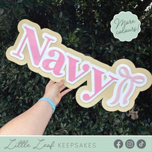 May include: A wooden sign spelling "Navy" in pink, with a white outline, and a light brown base. A pink bow is on the right. The sign is held against green leaves. A circular sign says "More colours!"