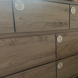 May include: A wooden chest of drawers with circular labels on each drawer. The labels read "Socks," "Hats," "Jumpers," and "Pyjamas," with matching illustrations. The chest has a light brown wood grain finish.