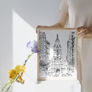 May include: A framed black and white ink drawing of a city skyline with a tall building in the center. The drawing is on a white background and is framed in a light wood frame.