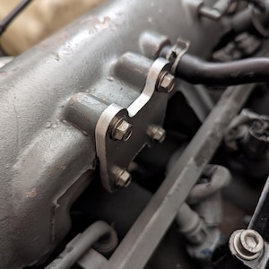 Può includere: Primo piano di un componente del motore con una staffa in metallo argentato fissata con diversi bulloni. La staffa è fissata a un blocco motore grigio strutturato, con tubi e tubi flessibili sullo sfondo.