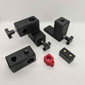 Pode incluir: Um conjunto de blocos de montagem retangulares pretos com botões de ajuste roscados, um pequeno componente vermelho em forma de L e um bloco retangular preto com duas inserções circulares douradas, concebidos para montagem mecânica.