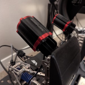 May include: Close-up of a black and red racing simulator pedal set. The pedals have a black footrest and a silver-colored metal frame. The cylinders have the word "SIMAGIC" printed on them.