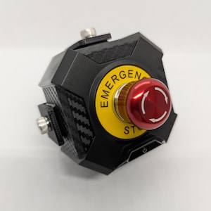 Könnte beinhalten: Ein schwarzer Not-Aus-Knopf aus Kohlefaser mit einem roten Druckknopf und einem gelben Etikett mit der Aufschrift "NOT-AUS".