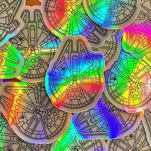 May include: A collection of holographic stickers featuring the Millennium Falcon spaceship from Star Wars. The stickers are silver with black outlines and have a rainbow iridescent finish.