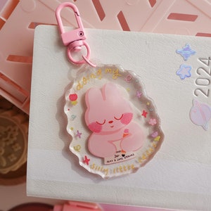 Peut inclure: Un porte-clés en acrylique rose avec un lapin de dessin animé tenant un cornet de glace. Le porte-clés a un fermoir rose et le texte "doing my silly little rabbit things that are cute". Il est posé sur un carnet blanc.